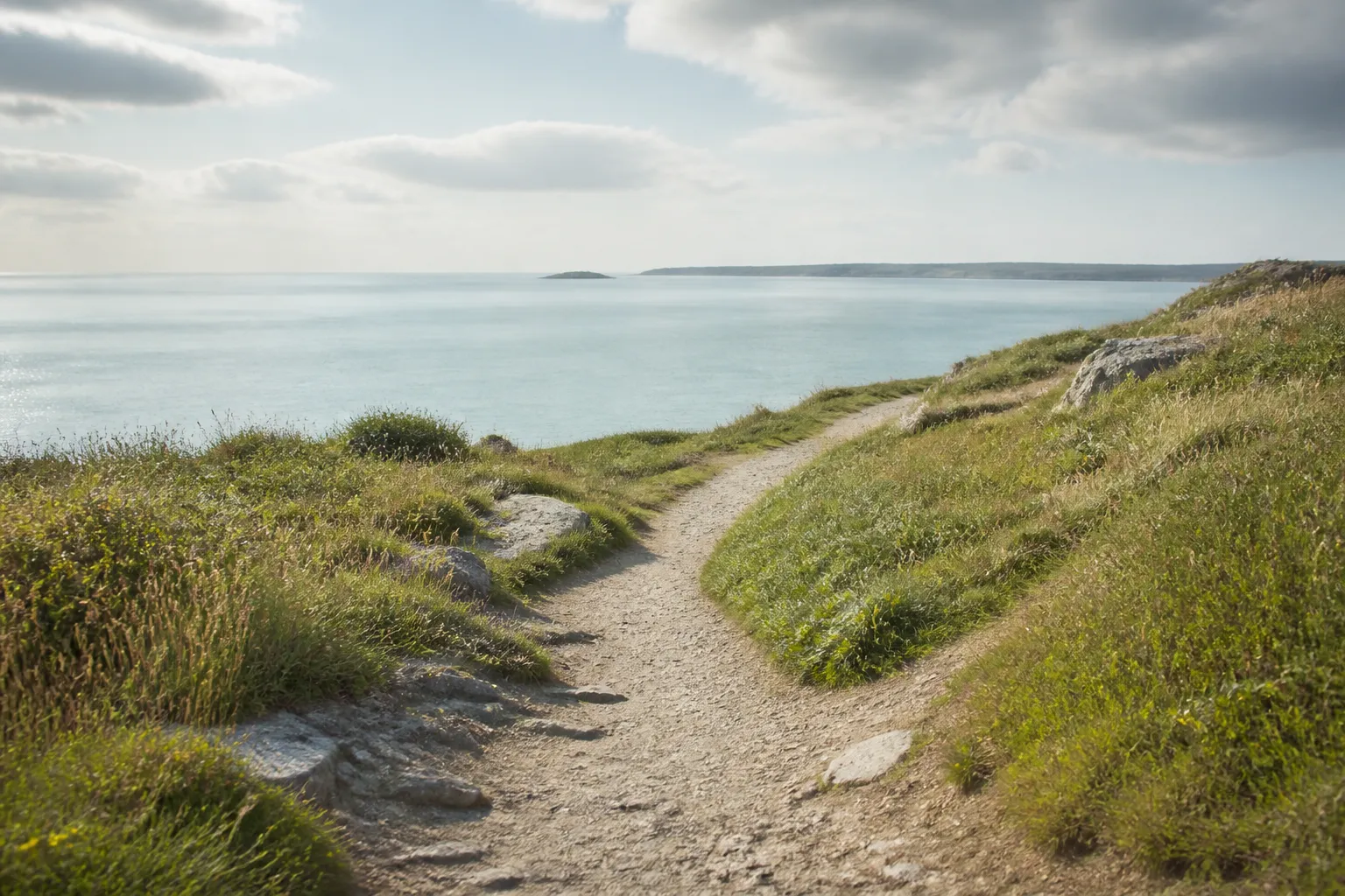 A quiet outdoor coastal path through grassy dunes towards calm pale water
